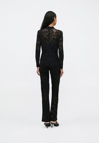 Salopetă neagră cu mâneci lungi, textură din dantelă florală transparentă, siluetă ajustată și picioare drepte. Include un guler înalt și închidere cu fermoar invizibil.