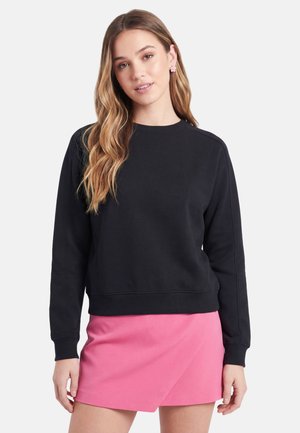 Junge Frau mit langen welligen Haaren, die einen schwarzen Sweatshirt und einen pinken Minirock trägt, steht vor einem schlichten hellen Hintergrund.
