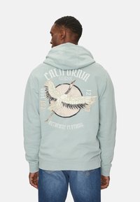 Sudadera de color verde menta con un gran gráfico de un pájaro y el texto "California" en la parte posterior. Cuenta con una capucha con cordones y dobladillos acanalados en el bajo y los puños.