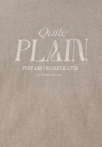 Väri bežissä kankaassa on tekstuuri, jossa on valkoista painettua tekstiä: "Aika YKSINKERTAISTA" ja "PUHDAS JA YKSINKERTAINEN RANNALLA" sen alapuolella.