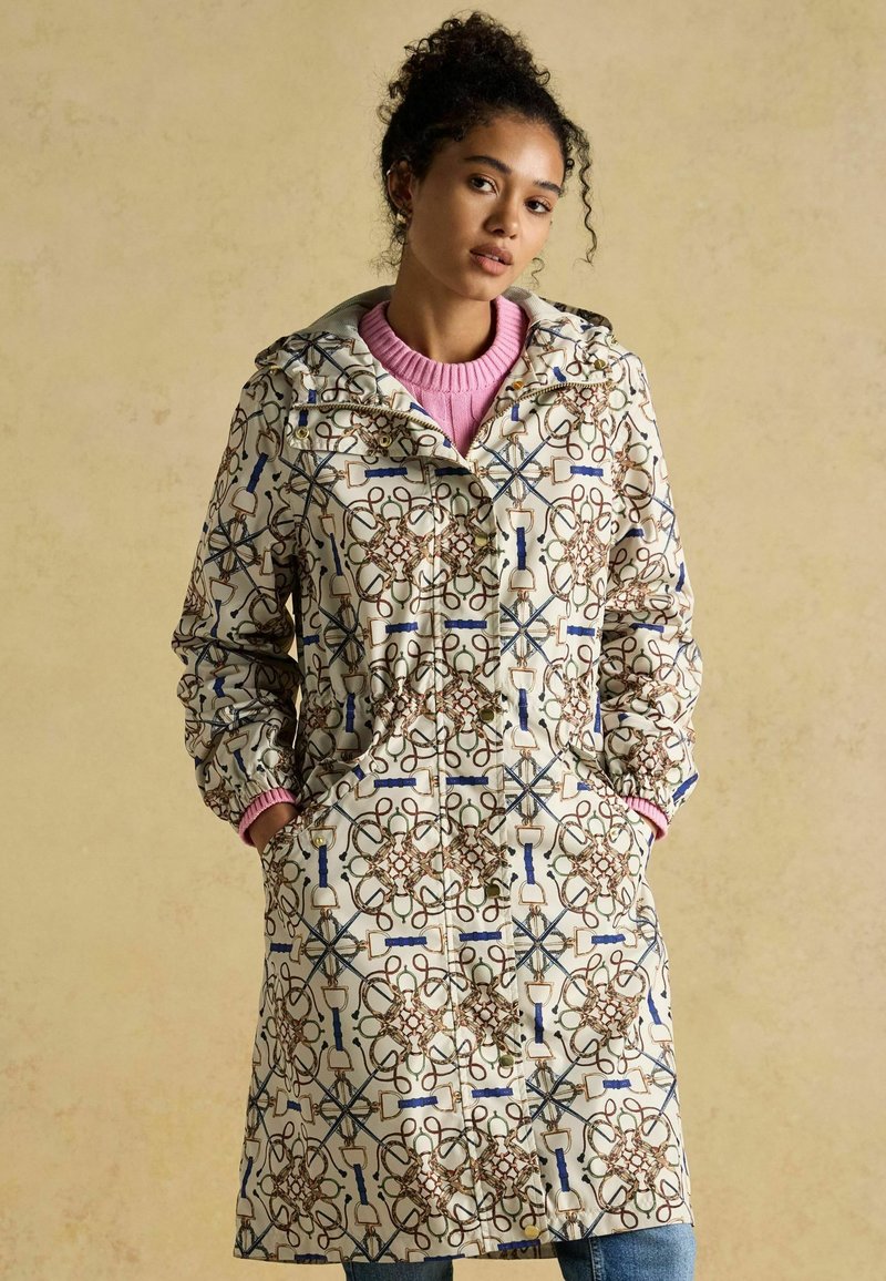 Sieviete valkā garu rakstainu mēteli ar kapuci, rokas kabatās, zem tā rozā džemperis, stāv uz vienkārša bēša fona.