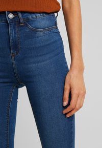 Vaqueros de mezclilla azules de tiro alto con un diseño clásico de cinco bolsillos, costuras sutiles y una textura suave, detalle del botón de la cinturilla visible.