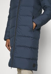 Jūras spēku puffer mētelis ar garu siluetu, horizontāli quiltēts dizains, divas sānu kabatas ar rāvējslēdzējiem un elastīgu piedurkņu apdari.
