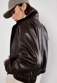 Braune Leder-Cropped-Jacke mit hohem Kragen, ausgestattet mit einem Reißverschluss vorne und einem elastischen Saum. Das Model trägt eine Hahnentritt-Mütze.