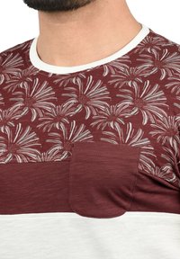 Burgundy t-shirt with floral print, white collar, horizontal color block design; features a front pocket and textured fabric.