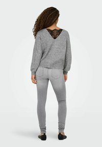 Pull gris avec un insert en dentelle dans le dos, manches longues et coupe loose, associé à un jean gris clair cintré et des chaussures noires plates.