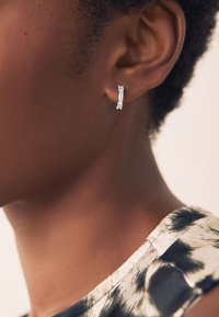 Silberne Ohrringe mit rechteckigen und runden klaren Steinen, die dicht beieinander gesetzt sind. Die braune Haut und das gemusterte Oberteil des Modells sind teilweise sichtbar.