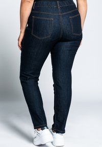 Jeans de mezclilla azul oscuro con una cinturilla elástica, dos bolsillos traseros y costuras en color naranja. Las piernas tienen un ajuste slim y se estrechan hacia el tobillo.