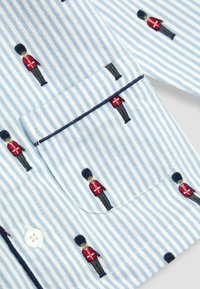 Svetlomodrá pruhovaná košeľa s malými vyšívanými vojakmi v červených kabátoch. Tlačidlový detail na vrecku na hrudi s námorníckym lemom. Mäkká bavlnená látka.