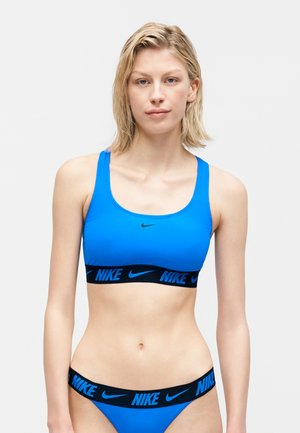 Kobieta w niebieskim sportowym biustonoszu Nike i dopasowanych majtkach z czarnymi elastycznymi paskami z logo Nike, stojąca na białym tle.