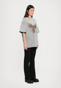Grijze oversized T-shirt met een bruine adelaarafbeelding. Korte mouwen met accentdetails. Gecombineerd met zwarte flare-broeken en gedurfde zwarte laarzen.