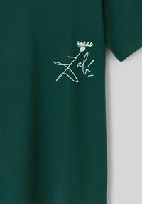 T-shirt vert foncé avec une signature stylisée blanche et un petit graphique de couronne sur la partie gauche de la poitrine.