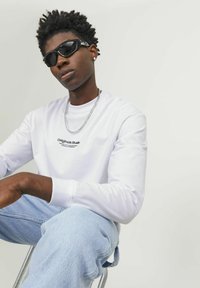 Long-sleeve white shirt with printed text, light blue denim jeans, and black sunglasses. Short black hair, wearing a silver chain necklace.