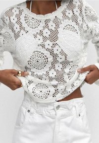 Bílý háčkovaný svetr s květinovými a kruhovými vzory, dlouhými rukávy a kulatým výstřihem, doplněný bílými kalhotami s vysokým pasem.