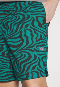 Shorts à motifs verts et noirs avec un design de vagues, fabriqués en tissu léger, avec une étiquette logo Dickies sur le côté.