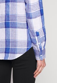 Camicia a quadri blu e bianca con maniche lunghe, polsini con bottoni e orlo curvo. Realizzata in tessuto leggero, che rivela pantaloni neri sottostanti.