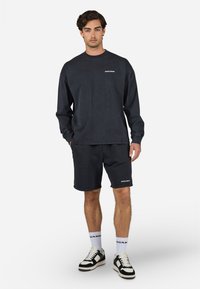 Maglietta a maniche lunghe grigio scuro e pantaloni coordinati con una stoffa testurizzata. Presenta il logo bianco "PECADOR". Sneakers in bianco e nero completano il look.