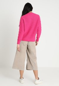Femme aux longs cheveux noirs portant un pull tricoté rose vif, un pantalon large beige coupé au-dessus de la cheville et des baskets blanches, debout devant un fond uni.
