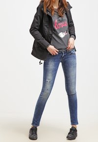 Giacca nera impermeabile con cappuccio, t-shirt grigia con testo rosso e bianco, jeans skinny blu consumati e sneakers nere.