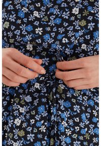 Květinové šaty s námořnickým pozadím, zdobené modrými, bílými a zelenými květinami. Detailní záběr rukou, které svazují látku pomocí uzlu.