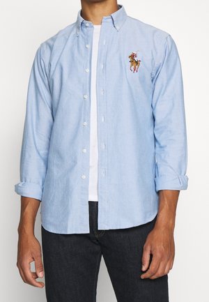 Mężczyzna noszący jasnoniebieską koszulę na guziki na białej koszulce, koszula posiada haftowane logo gracza polo, rękawy podwinięte, zestawiona z ciemnymi dżinsami.