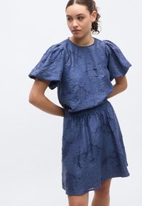 Robe bleue texturée avec manches bouffantes, taille élastique et ourlet au niveau des genoux. Le tissu présente un motif froissé, offrant un détail visuel unique.