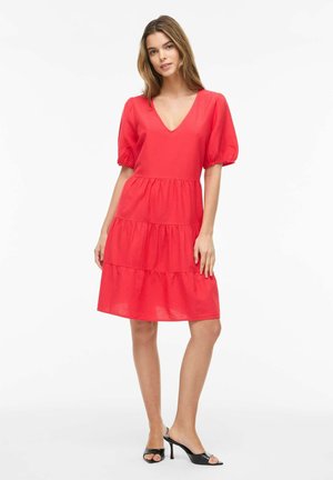 Žena v červenom šatách po kolená s nadýchanými krátkymi rukávmi a výstrihom do V, stojaca s jednou rukou na boku, obutá v čiernych lodičkách na vysokom podpätku.
