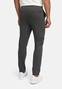 Man wearing dark gray slim-fit trousers and white sneakers, shown from the back against a plain light background.