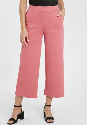 Pantalons à large jambe rose en tissu doux, avec une taille haute et des poches latérales. Associés à des sandales à talons noires.