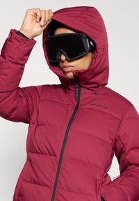 Veste rembourrée marron avec capuche, présentant des sections matelassées et une fermeture éclair noire. Des lunettes de ski noires couvrent les yeux, renforçant la fonctionnalité en extérieur.