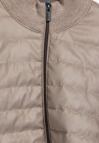 Beige gewatteerde jas met kleine stippenstructuur, zwarte rits en geribbelde kraagdetail.