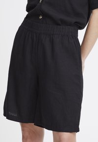 Shorts en lin noirs avec une taille élastique, de longueur genou, présentant une coupe décontractée et des poches latérales. Le tissu semble léger et texturé.