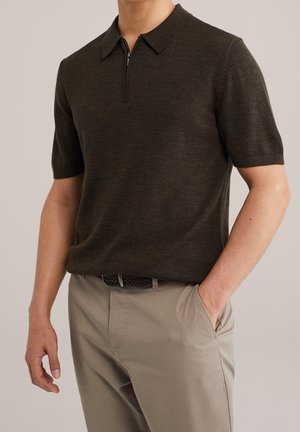 Mand iført mørkebrun kortærmet sweater med lynlåskrave og beige bukser med flettet sort bælte, den ene hånd i lommen mod ensfarvet baggrund.