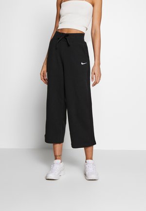 Woman wearing black Nike wide-leg cropped pants, white cropped tank top, and white sneakers, standing against a plain background.