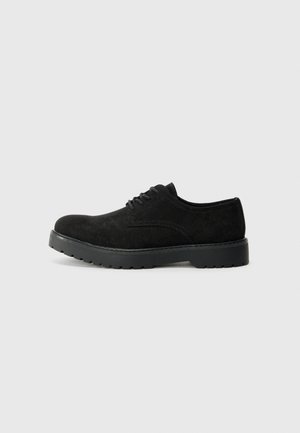 Chaussure en suède noir à lacets avec un bout arrondi, tige texturée et semelle en caoutchouc noir solide présentant un motif de semelle.