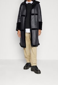 Abrigo de cuero negro con acentos de piel, con un frente abotonado, bolsillos tipo parche y una capucha con cordón. Lleva puesto un suéter oscuro y pantalones beige.