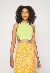 Top cropped verde com textura canelada, apresentando recortes laterais e acessórios prateados. Combinado com uma saia laranja fluida com padrão em espiral.