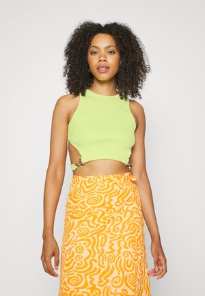 Top cropped verde com textura canelada, apresentando recortes laterais e acessórios prateados. Combinado com uma saia laranja fluida com padrão em espiral.