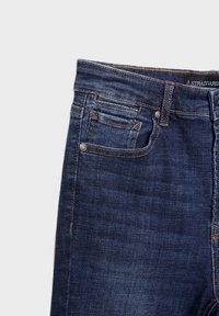 Jean en denim bleu foncé avec un design classique à cinq poches, présentant des coutures orange contrastantes et une petite étiquette logo sur la taille.
