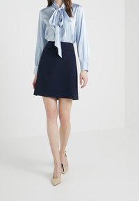 Light blue satin blouse with bow tie collar, long sleeves. Dark navy A-line skirt with structured texture. Beige pointed-toe heels complete the look.
