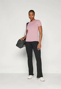 Naine seisab tasasel taustal, kandes roosa lühikeste varrukatega särki, musti pükse, valgeid tossusid ja hoides musta õlakotti.