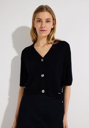 Blonde vrouw die een zwart gebreid vest met korte mouwen en drie knopen draagt, staand tegen een effen lichte achtergrond.