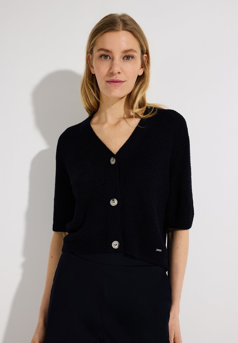 Blonde vrouw die een zwart gebreid vest met korte mouwen en drie knopen draagt, staand tegen een effen lichte achtergrond.