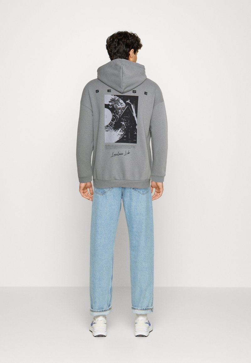 Homme portant un sweat à capuche gris avec un graphisme et un texte dans le dos, un jean bleu clair et des baskets blanches, debout face à un mur uni.