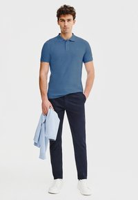 Niebieska koszulka polo z krótkim rękawem, gładka tekstura, z kołnierzykiem. W połączeniu z granatowymi spodniami i trzymana jasnoniebieska bluza. Białe buty.