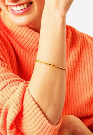 Bracciale in metallo dorato con un design intrecciato, caratterizzato da una superficie liscia. Indossato su un polso nudo, in contrasto con un maglione coral texture.
