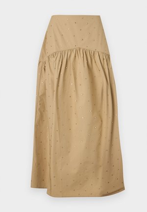 Donna con capelli lunghi indossa una giacca corta beige con bottoni dorati, un top bianco senza maniche, una gonna ampia color cuoio con piccoli motivi e décolleté neri.