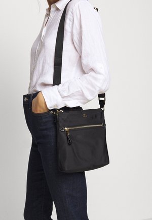 Personne portant une chemise blanche à manches longues et un jean foncé, portant un sac bandoulière noir avec une poche avant zippée et une sangle d'épaule réglable.