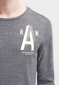 Harmaa pitkähihainen paita, jossa on suuri valkoinen ja metallinhohtoinen harmaan "A" -grafiikka, ja sen alla teksti "PURVEYORS OF 3D DENIM". Sujuva pinta.