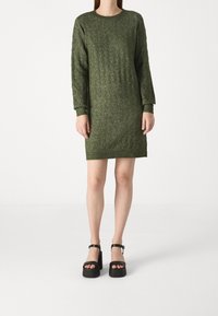 Abito in maglia verde scuro con un motivo testurizzato, maniche lunghe e scollatura rotonda. Abbinato a sandali con plateau neri con cinturini alla caviglia.